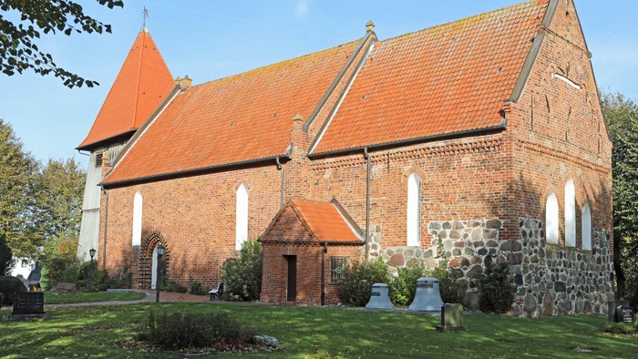 Das Bild zeigt eine rote Backsteinkirche mit spitzen Dächern und weißen Fenstern, umgeben von grünem Gras und Bäumen.