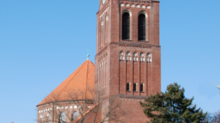 Große rote Backsteinkirche mit hohem Turm und spitzem Dach, umgeben von Bäumen und einem klaren blauen Himmel