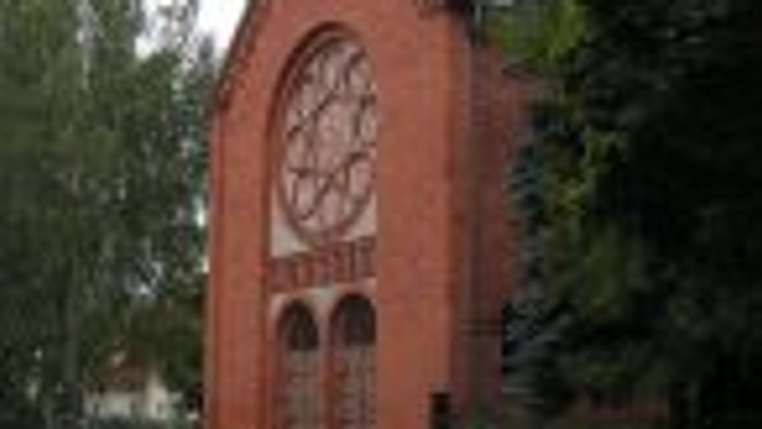 Eine rote Backsteinkirche mit Bogenfenstern und einem hohen Kirchturm, umgeben von üppigen grünen Bäumen.