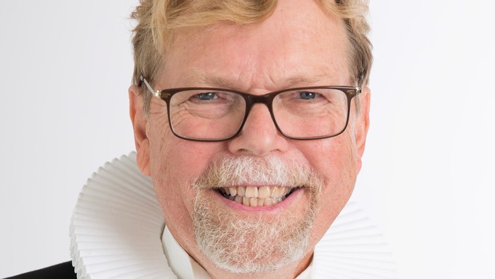 En smilende mand med blond hår og bart, der bærer en sort jakke med hvid krage.