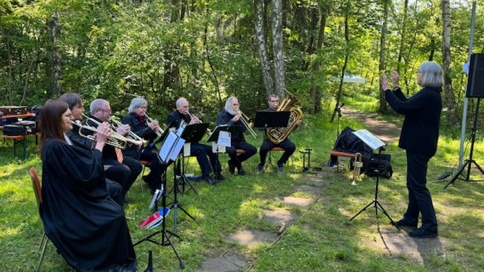 Draußen musizierendes Blechbläserensemble unter Bäumen mit einem Dirigenten, der die Musikprobe leitet.