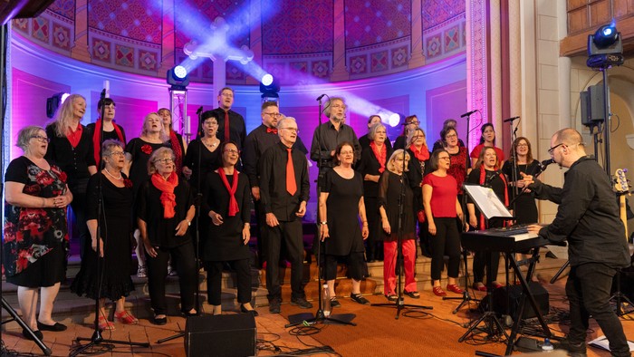 Ein Chor tritt auf einer Bühne auf, dirigiert von einem Mann am Klavier