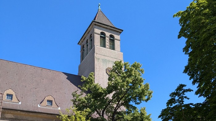 Kirchturm mit Uhr und Kreuz, umgeben von Bäumen und Gebäuden