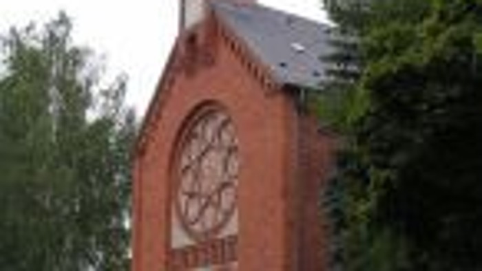 Eine rote Backsteinkirche mit Bogenfenstern und einem hohen Kirchturm, umgeben von üppigen grünen Bäumen.