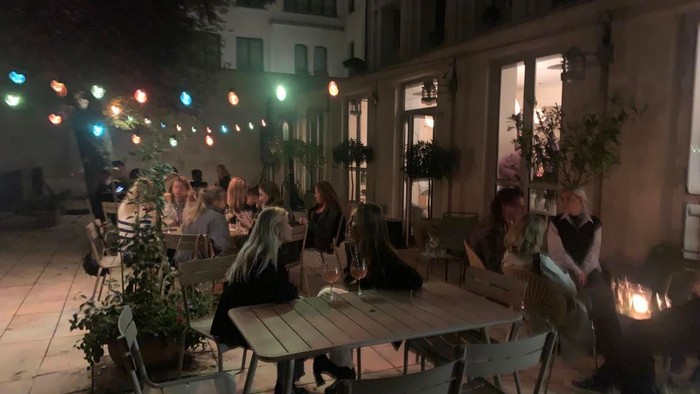 En ude spisestue med folk der sidder ved bord og taler, belysning fra stråler og planter.