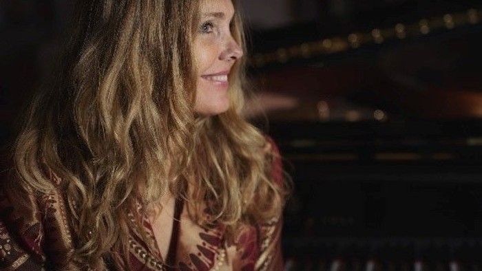 En kvinde med lang, blond hår sitter ved et piano i en koncertsal.