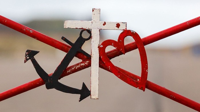 Ein schwarzes Anker-Symbol hängt an einem weißen Kreuz, das an einem roten Metallgitter befestigt ist, daneben ein rotes Herz-Symbol