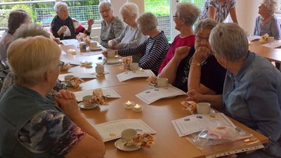 En gruppe pensionister sidder ved bord og spiser kage og drikker kaffe.