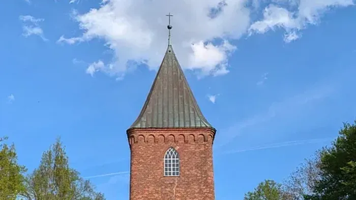 Kirchturm aus rotem Backstein vor blauem Himmel und Bäumen