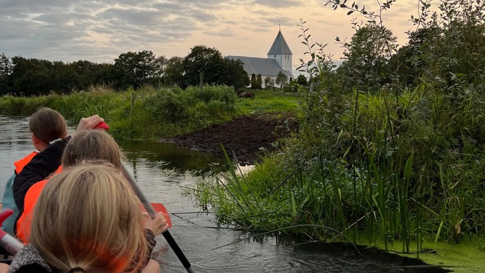 Piger med orange redningsveste på, der ror i kano på en å. I baggrunden Nr. Løgum kirke under er aftenhimmel med lyse skyer