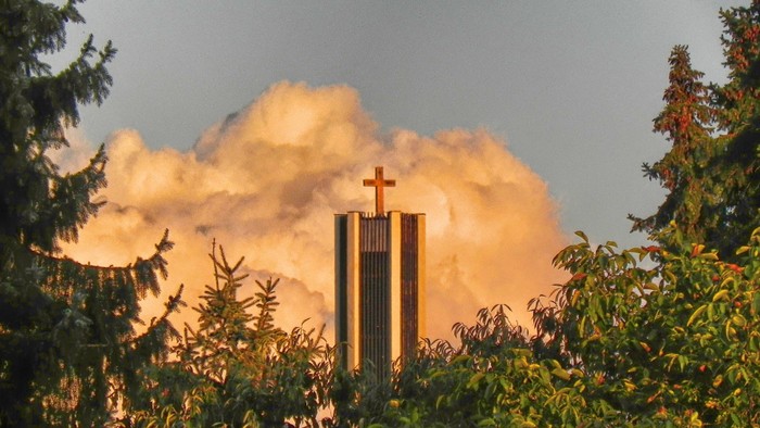 Ein hoher Turm mit Kreuz vor Sonnenuntergang und Wolken, umgeben von Bäumen