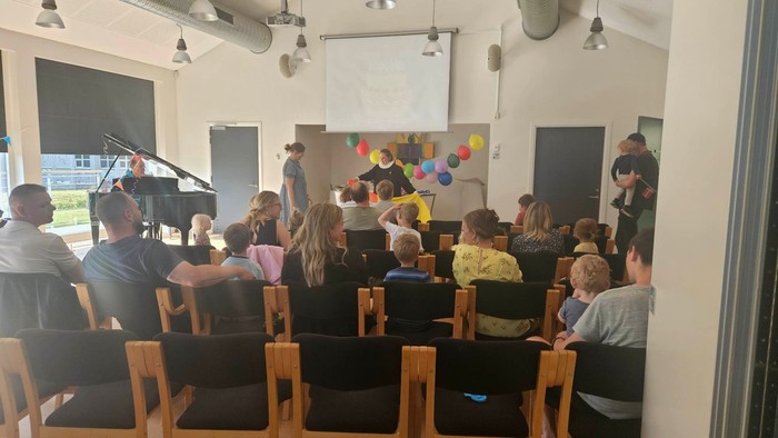 En gruppe børn og voksne sidder i en hall og ser en foredragsholder.