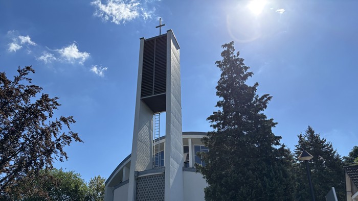 Moderne Kirche mit hohem Glockenturm vor strahlend blauem Himmel und umgeben von Grün.