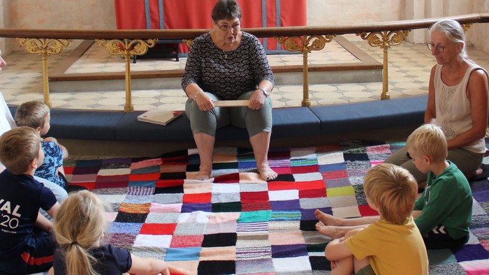 En flok børn sidder på gulvet i en halvcirkel og lytter til en fortæller. Fortælleren sidder på knæfaldet i Nr. Løgum kirke