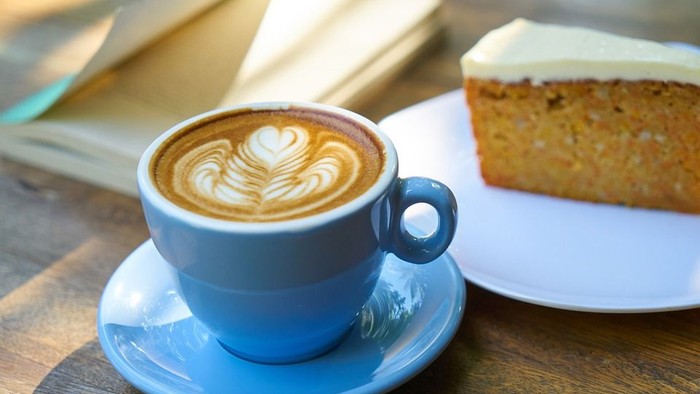 Ein blauer Kaffeebecher mit Milchschaum und ein Stück Kuchen auf einem Holztisch