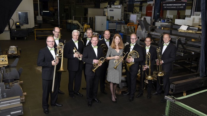 Eine formelle Blaskapelle posiert gemeinsam in einer industriellen Werkstatteinrichtung mit verschiedenen Maschinen.