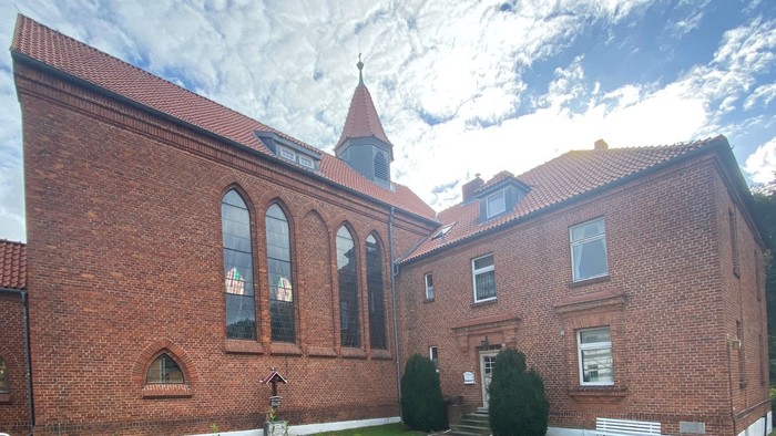 Ein rotes Backsteingebäude mit spitzem Turm und bogenförmigen Fenstern unter einem bewölkten Himmel.