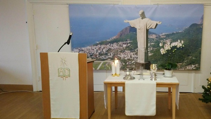 Ein Tisch mit weißen Tischtuch, Kerzen und einem Kruzifix davor, vor einem Bild von Christus der Erlöser in Rio de Janeiro