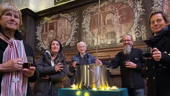 Eine Gruppe von Menschen steht um einen Tisch mit einem großen Pott, alle lächeln und halten Getränke.