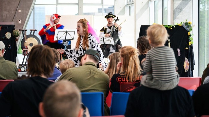 En gruppe børn og voksne ser en show med to pupper og en mand spillende violin.