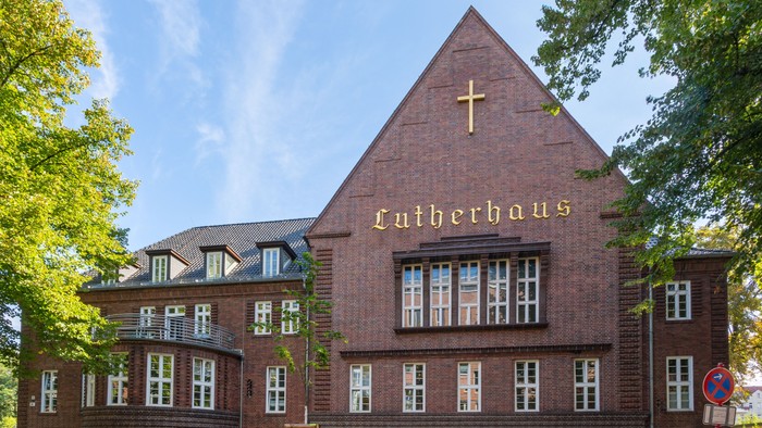Das Bild zeigt ein großes, rotes Gebäude mit einem Kreuz und der Aufschrift 