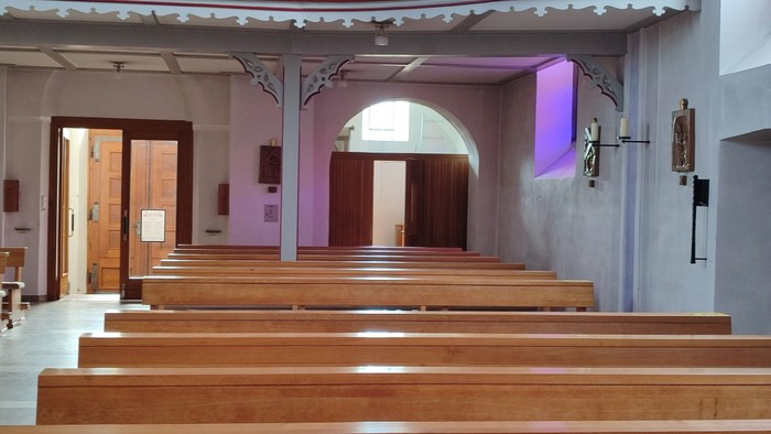 Leerer Kircheninnenraum mit hölzernen Kirchenbänken, die zu einem gewölbten Altarraum unter sanftem violettem Licht führen.