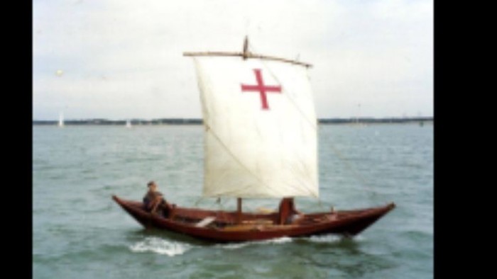A small boat with a white sail marked with a red cross sails on calm waters.