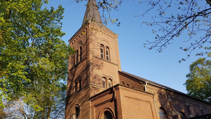 Das Bild zeigt einen hohen Kirchturm aus Ziegeln vor grünen Bäumen und blauem Himmel