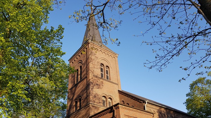 Das Bild zeigt einen hohen Kirchturm aus Ziegeln vor grünen Bäumen und blauem Himmel