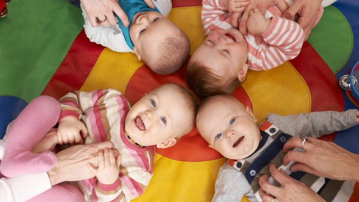 Fünf Babys liegen im Kreis auf einer bunten Decke und werden von Erwachsenen gehalten.