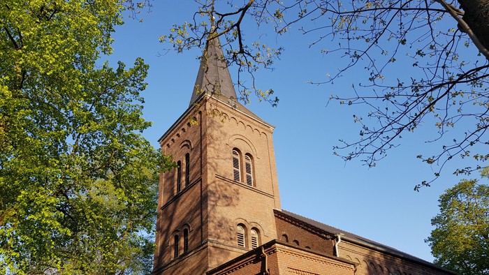 Das Bild zeigt einen hohen Kirchturm aus Ziegeln vor grünen Bäumen und blauem Himmel