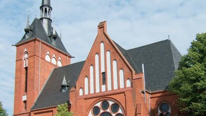 Historische Backsteinkirche mit hohem Turmspitz und rundem Rosenfenster unter einem teilweise bewölkten Himmel.