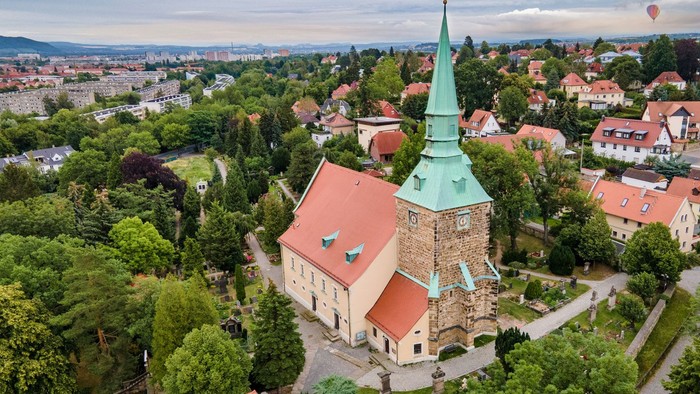 Das Bild zeigt eine Kirche mit hohem Turm in einem grünen, dörflichen Umfeld.