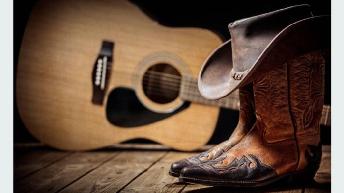 Cowboy boot next to an acoustic guitar on wooden floor
