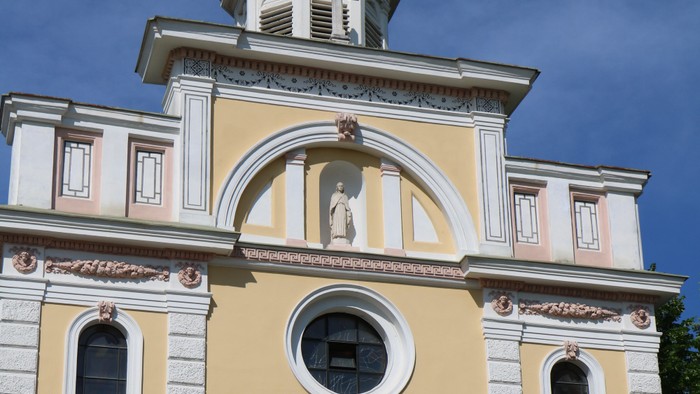 Das Bild zeigt eine Kirche mit gelben Wänden und weißem Zierwerk, einem Uhrturm und einer Statue.