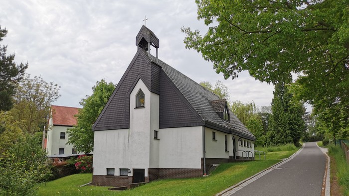 Kleine Kirche mit schwarzem Dach und weißer Fassade, umgeben von Bäumen und einem Weg.