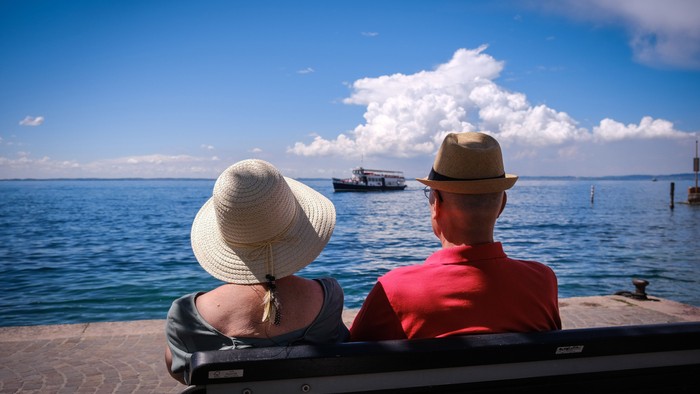 Ein Paar sitzt auf einer Terrasse am Meer und schaut auf ein Boot.