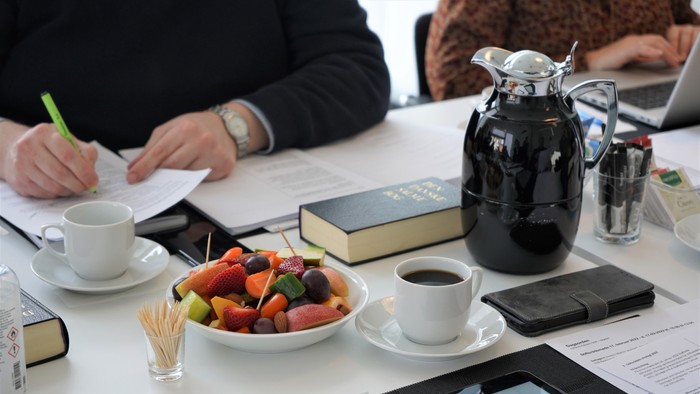 To dagslys og en gruppe af mennesker sidder ved et bord med notatblokke, kaffe, frukt og en bible.