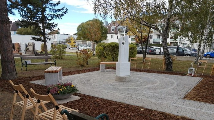 Ein kleiner Park mit weißem Denkmal, Bänken und Blumenbeeten.