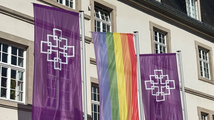 Vor einem Gebäude hängen drei Fahnen: zwei lila mit weißem Kreuz und eine Regenbogenfahne.
