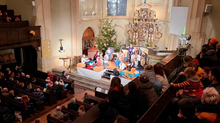 In der Kirche feiern viele Menschen Weihnachten, ein geschmückter Baum und ein Altar sind zu sehen.