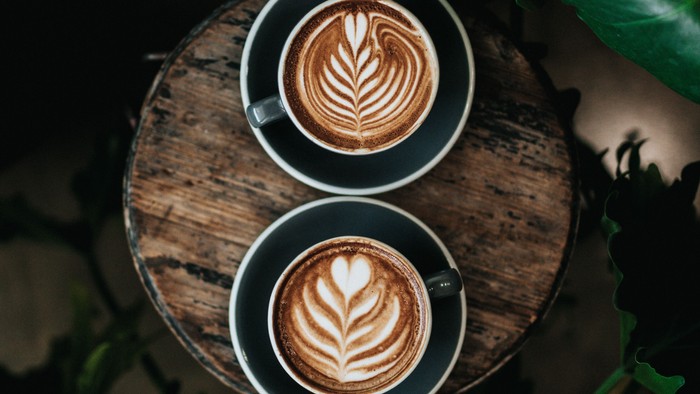 Zwei Kaffeetassen auf Holztisch mit Blattmuster, umgeben von Grünpflanzen