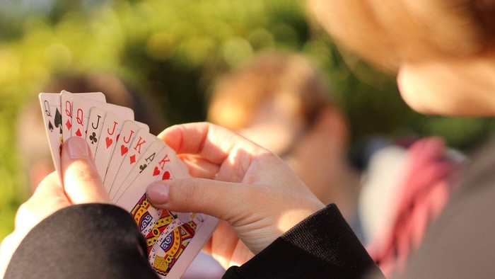 Eine Hand hält einen Fächer aus Spielkarten, im Hintergrund sitzt jemand im Freien.