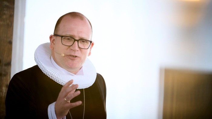 En person i formelt tøj med en krusekrave og briller ser ud til at tale eller forklare noget.