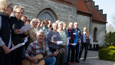 En gruppe af mennesker står foran en kirke, hvor nogle spiller guitar og andre holder noter.