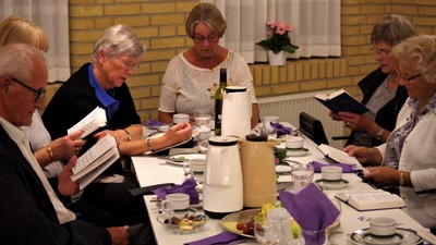 En gruppe ældre personer sidder ved et bord med mad og drikkevarer