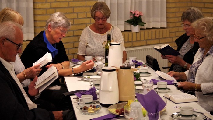 En gruppe ældre personer sidder ved et bord med mad og drikkevarer