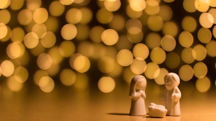 Two small figurines stand beside a manger, with a blurred background of golden lights.