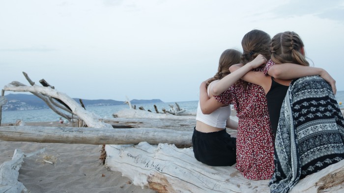 Drei Personen sitzen auf Treibholz am Strand, umarmen sich und blicken auf das Meer hinaus.