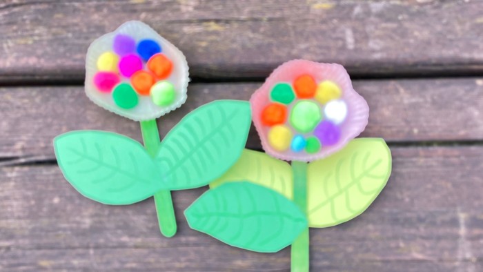 Die Abbildung zeigt eine Nahaufnahme von zwei gebastelten Blumen auf einem Hintergrund aus Holz. Die Blumen bestehen aus grünem Papier für die Blätter und grünen Holzspateln für die Stiele. Die Blumenköpfe sind Muffinförmchen mit bunten Pompons beklebt.